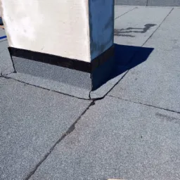 Wykończenie dachu papą wokół komina z użyciem obróbek blacharskich, widoczna listwa pomiarowa firmy Karl Rolli, na dachu widoczne łączenia papy.