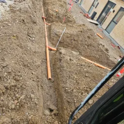 Wykop z pomarańczowymi rurami kanalizacyjnymi ułożonymi w ziemi, widoczne zabezpieczenia wykopu i fragment elewacji budynku w tle.