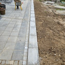Mat- bruk Mateusz Buschan - Fragment terenu z ułożoną szarą kostką brukową, obrzeżem z jasnych kamieni i przygotowanym podłożem z ziemi. W tle taczka z ziemią i worki z materiałem.