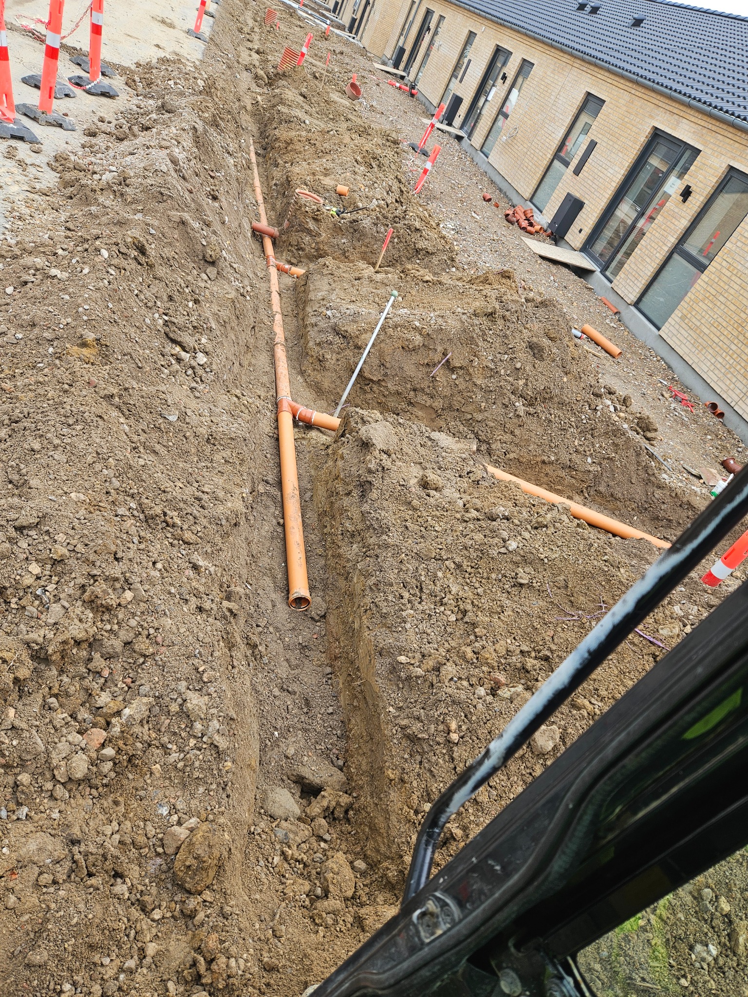 Wykop z pomarańczowymi rurami kanalizacyjnymi ułożonymi w ziemi, widoczne zabezpieczenia wykopu i fragment elewacji budynku w tle.