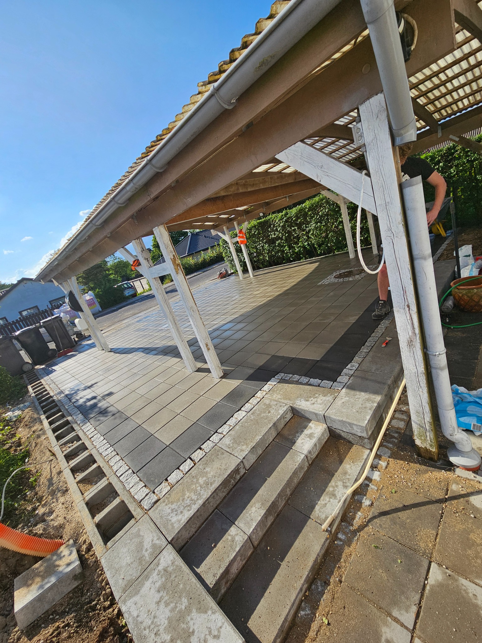 Zadaszona wiata z nową, szarą kostką brukową ułożoną w regularny wzór, obrzeża wykończone kostką granitową, widoczne schody wejściowe i przygotowane podłoże pod dalsze prace brukarskie.