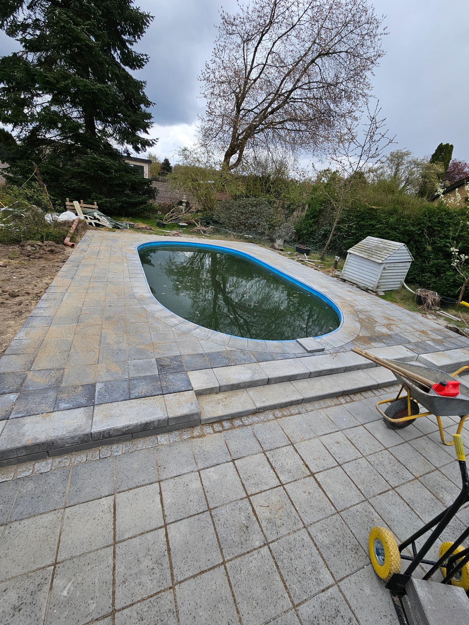 Owalny basen otoczony nowo ułożoną, szarą kostką brukową, widoczne schodki z kostki i taczka z narzędziami, w tle zieleń ogrodu i pochmurne niebo.