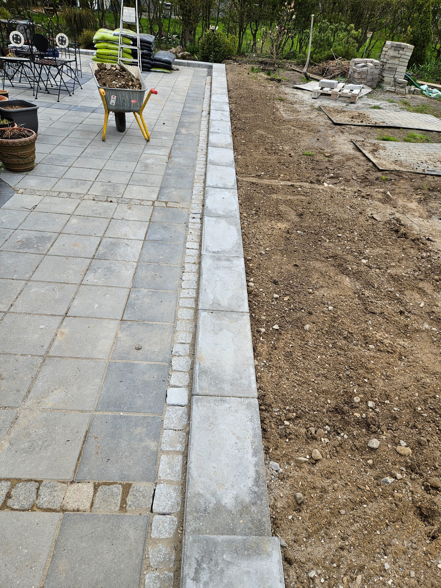 Fragment terenu z ułożoną szarą kostką brukową, obrzeżem z jasnych kamieni i przygotowanym podłożem z ziemi. W tle taczka z ziemią i worki z materiałem.