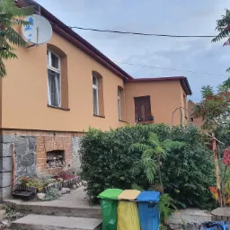 Dom z ocieploną elewacją w odcieniach beżu, z widoczną anteną satelitarną zabezpieczoną folią i taśmą, oraz fragmentem kamiennej podmurówki.