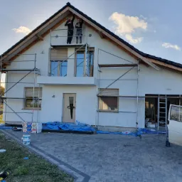 Dwóch pracowników na rusztowaniu przy dwukondygnacyjnym domu jednorodzinnym z jasną elewacją w trakcie prac wykończeniowych przy dachu, widoczne materiały budowlane i samochód dostawczy na posesji.