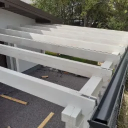 Konstrukcja drewniana z białych belek pod nowy dach, widoczne mocowania metalowe, rynna w kolorze grafitowym, w tle zieleń drzew.