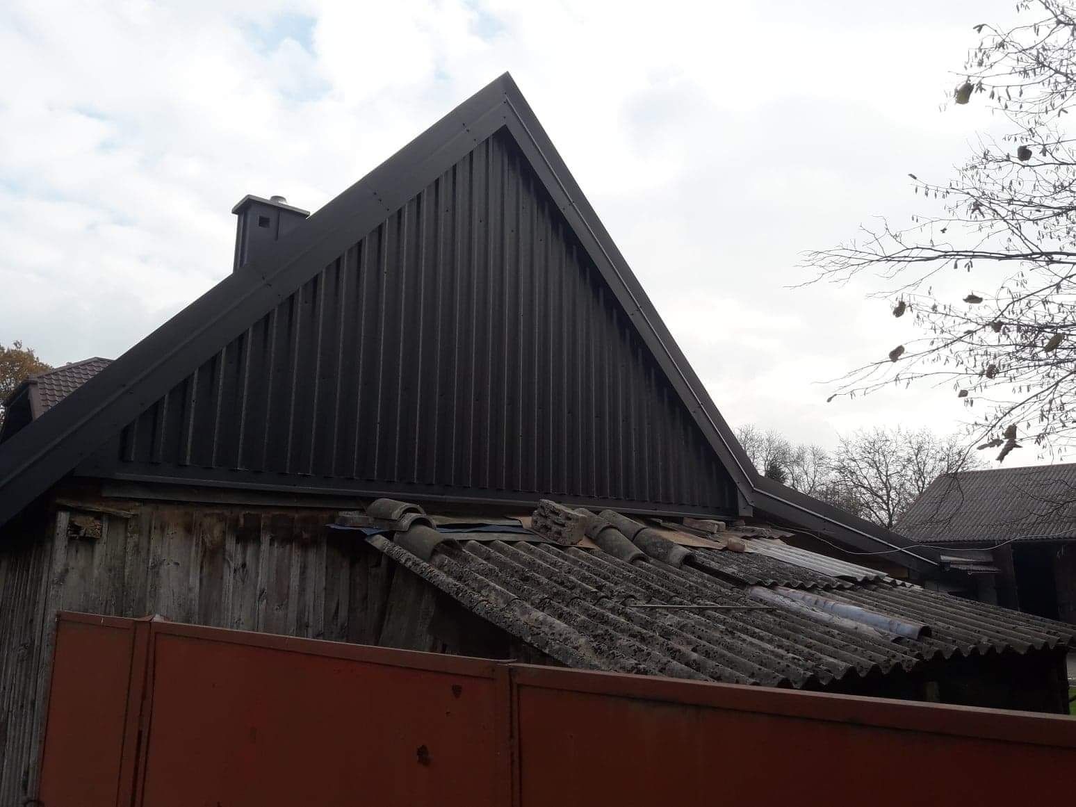 Ciemna, metalowa konstrukcja dachu z kominem, przylegająca do drewnianej ściany budynku, z fragmentem starego, zniszczonego dachu pokrytego eternitem w tle i fragmentem czerwonego ogrodzenia...