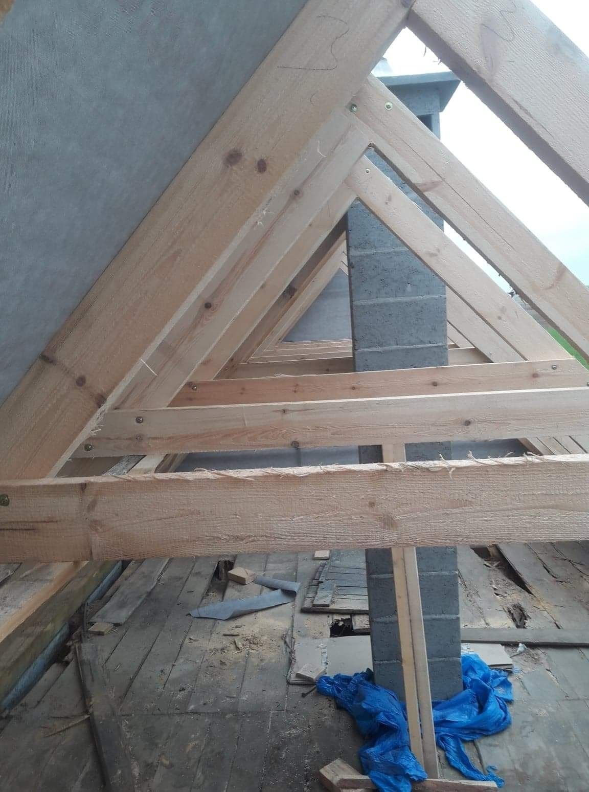 Konstrukcja drewniana dachu w trakcie budowy, widoczne krokwie, murłatki i komin z bloczków betonowych, zabezpieczone folią budowlaną, na podłodze resztki materiałów budowlanych i niebieska folia.