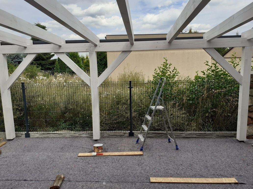 Drewniana konstrukcja z białymi belkami, prawdopodobnie zadaszenie tarasu, w trakcie malowania, widoczna drabina, puszka farby i deski na szarej macie zabezpieczającej podłoże.