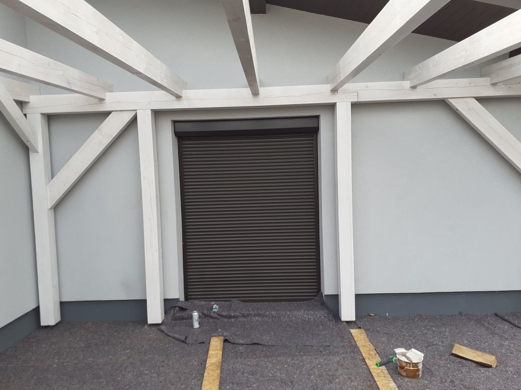 Zewnętrzna roleta w kolorze antracytowym, zamontowana pod drewnianą konstrukcją pergoli z belek pomalowanych na biało, widoczne ślady prac wykończeniowych na podłożu.