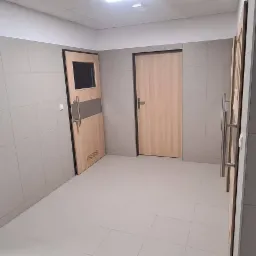 Korytarz wyłożony szarymi, kwadratowymi płytkami z widocznymi dwoma drzwiami z jasnego drewna, jeden z pionowym, ciemniejszym pasem i szybką, oświetlenie sufitowe