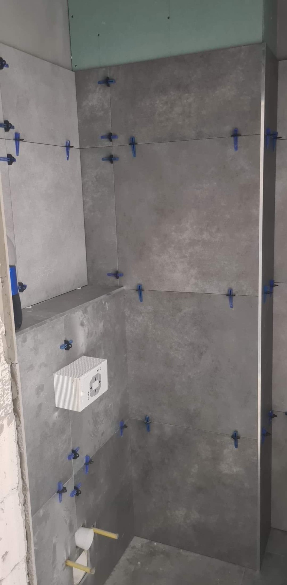 Szare płytki imitujące beton na ścianach łazienki, wyrównane niebieskimi klipsami poziomującymi, widoczna instalacja podtynkowego WC i fragment płyty gipsowo-kartonowej na górze ściany.