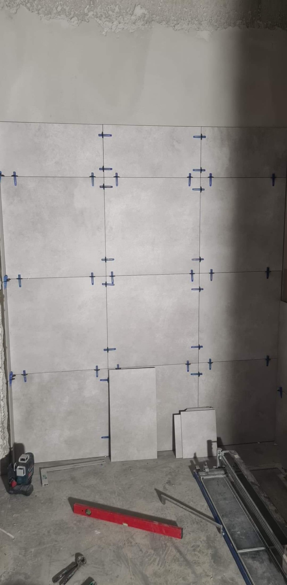 Montaż szarych płytek wielkoformatowych na ścianie z użyciem poziomicy laserowej i systemu poziomowania, widoczne narzędzia glazurnicze.