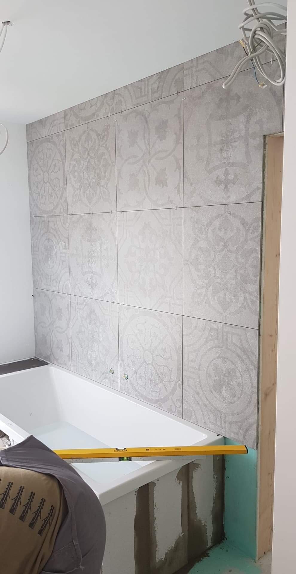 Ściana w łazience w trakcie układania płytek z delikatnym wzorem, poziomica oparta o wannę, widoczne przewody elektryczne.