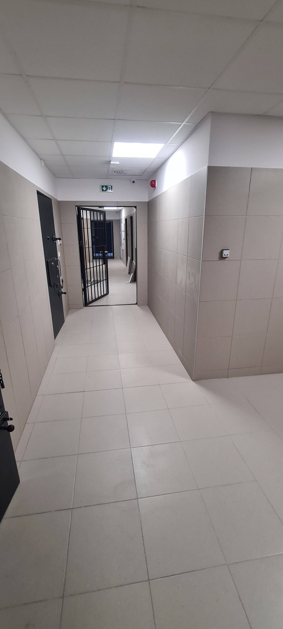 Korytarz wyłożony jasnymi, kwadratowymi płytkami podłogowymi i częściowo płytkami ściennymi, z otwartymi, metalowymi kratami w głębi i czarnymi drzwiami po lewej stronie, oświetlony równomiernym...