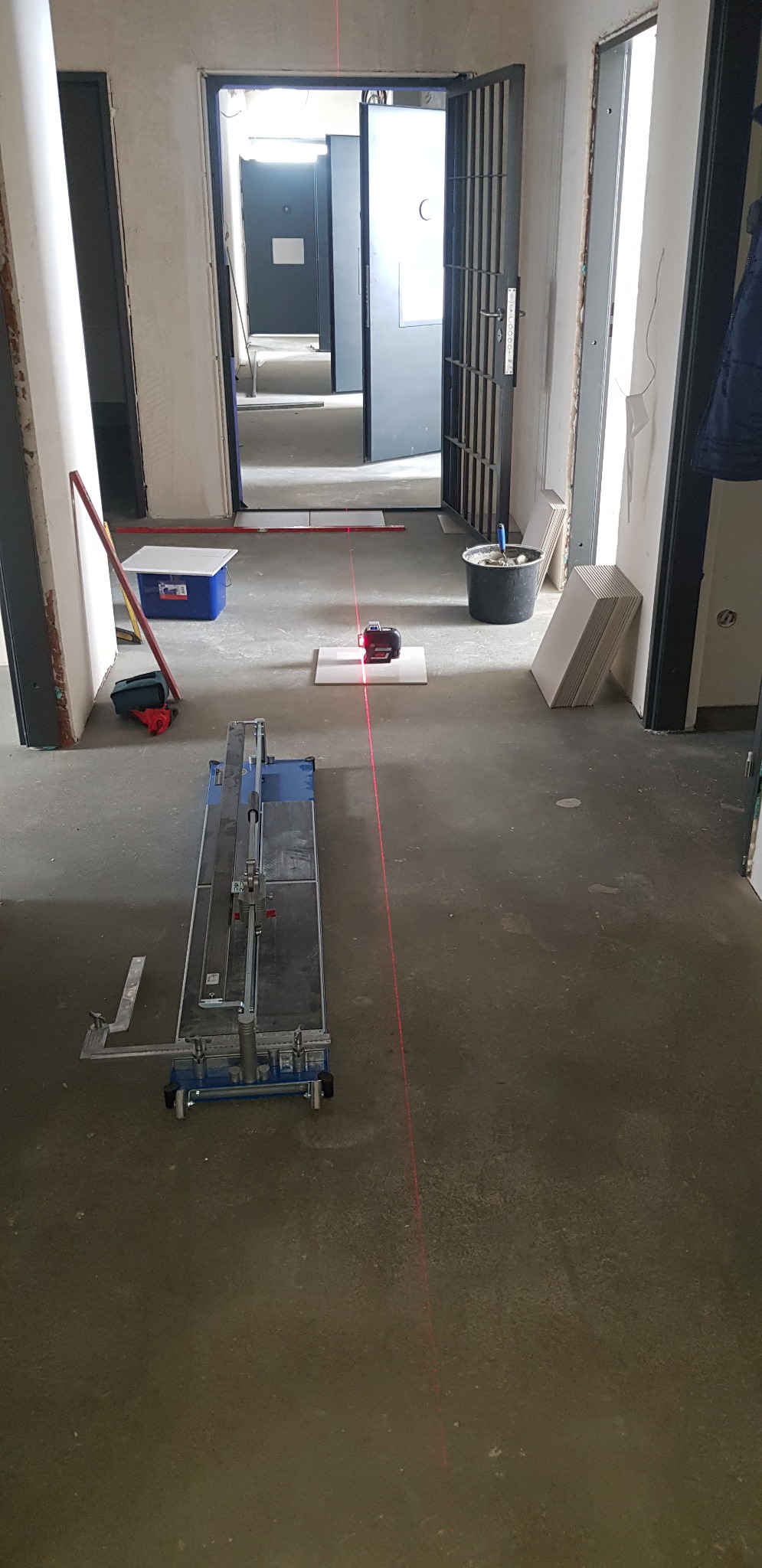 Przygotowanie do układania płytek w korytarzu: laser krzyżowy wyznacza linię, w tle otwarte drzwi i narzędzia glazurnicze.