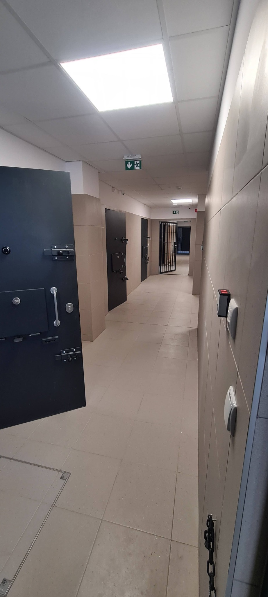Korytarz w Nowym Sączu z beżowymi płytkami na ścianach i podłodze, czarne metalowe drzwi z zamkami i łańcuchami, oświetlenie sufitowe i znaki ewakuacyjne.