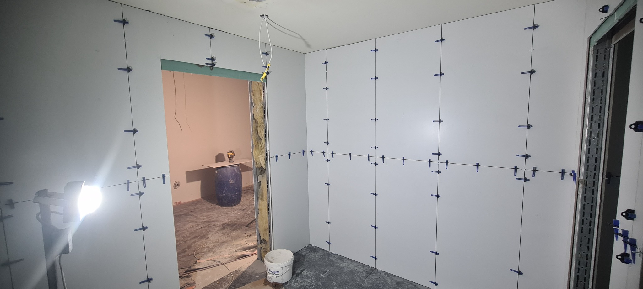 Przygotowanie ścian do położenia płytek w łazience w Nowym Sączu. Ściany wyłożone płytami kartonowo-gipsowymi, z widocznymi niebieskimi klipsami poziomującymi. W tle widoczne drzwi do innego...