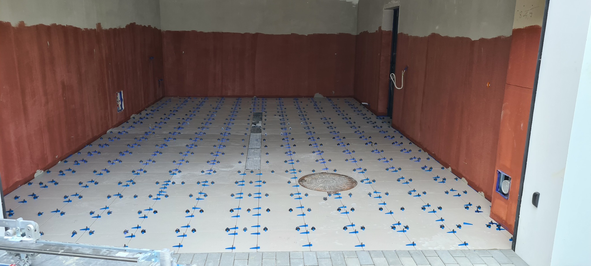 Jasne płytki podłogowe w trakcie układania, z niebieskimi krzyżykami dystansowymi, widoczny odpływ liniowy i studzienka rewizyjna, ściany częściowo przygotowane pod dalsze prace.