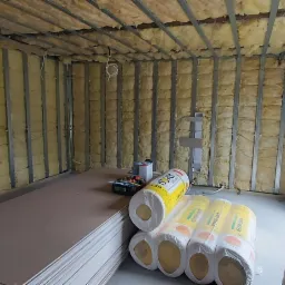 Wnętrze pokoju w trakcie prac wykończeniowych: ściany i sufit z izolacją z wełny mineralnej, zamontowane profile stalowe, stos płyt kartonowo-gipsowych oraz rolki wełny mineralnej na podłodze...