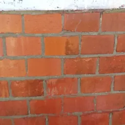 Fragment muru z czerwonej cegły, częściowo pomalowany ciemniejszą farbą, z widocznymi śladami zacieków i niedokładnego krycia.