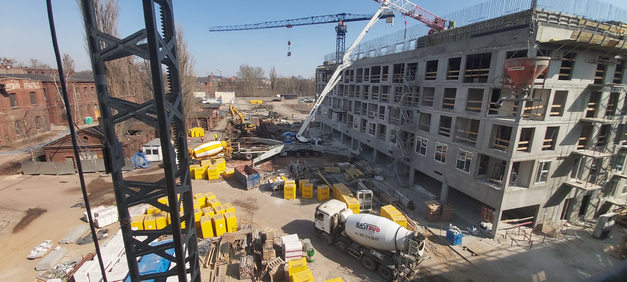 Wysoki kąt widzenia na plac budowy z betoniarkami, dźwigami i budynkiem w stanie surowym; widoczne materiały izolacyjne w żółtych opakowaniach.