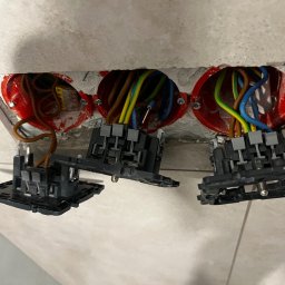 Mateusz Parchimowicz MIMAX - Potrójne gniazdko elektryczne w trakcie montażu, widoczne przewody w kolorowych izolacjach i puszki instalacyjne w ścianie wyłożonej płytkami.