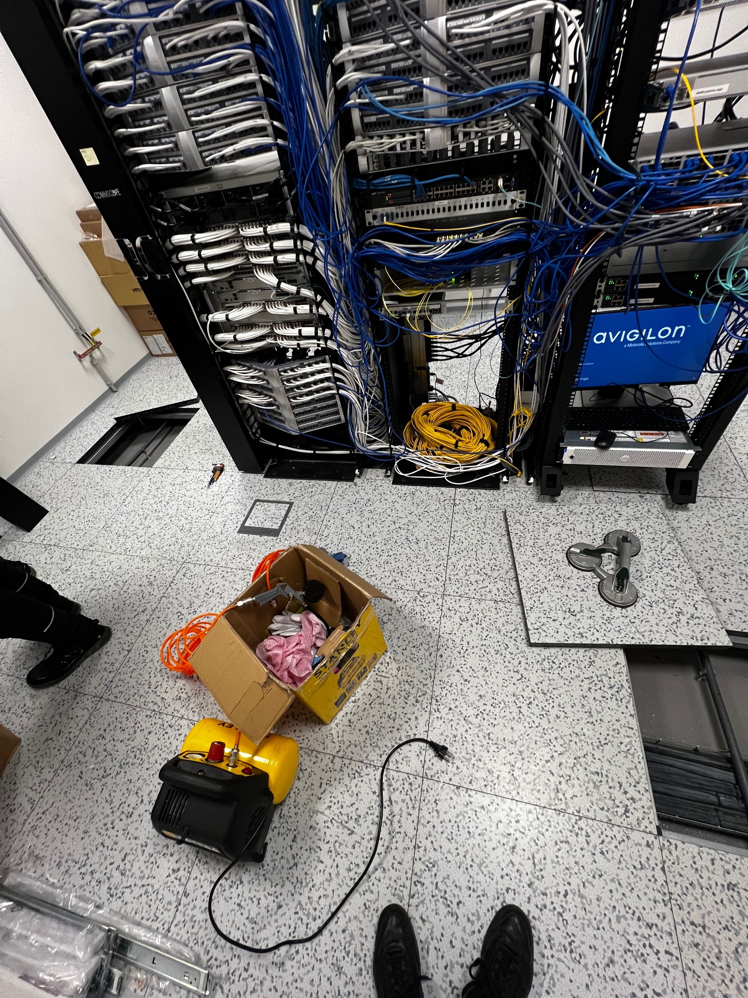 Serwerownia w trakcie instalacji: szafa rack z okablowaniem, narzędzia w kartonie, podniesiona podłoga techniczna. Widoczne kable, przełączniki i sprzęt Avigilon.