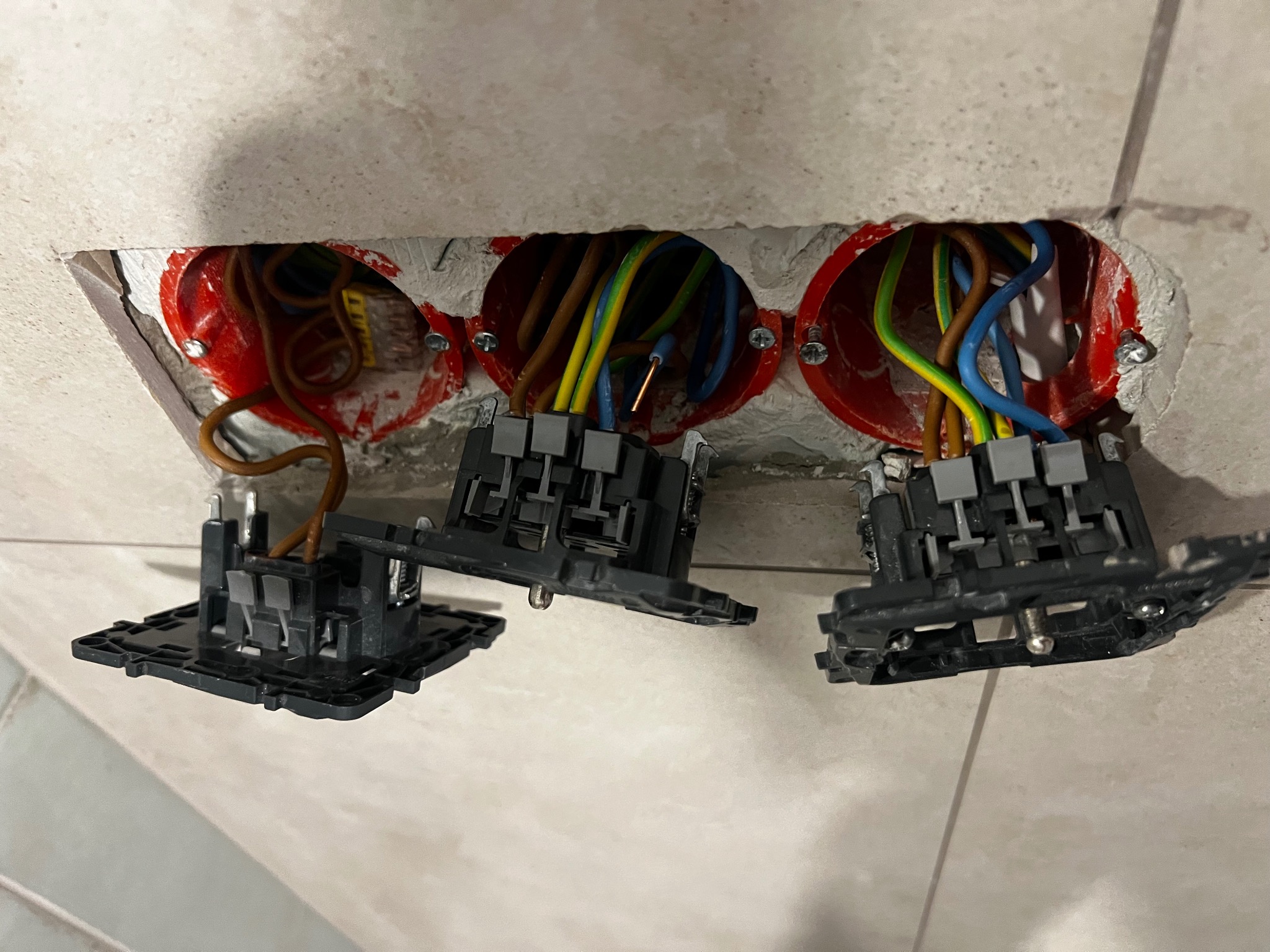 Potrójne gniazdko elektryczne w trakcie montażu, widoczne przewody w kolorowych izolacjach i puszki instalacyjne w ścianie wyłożonej płytkami.