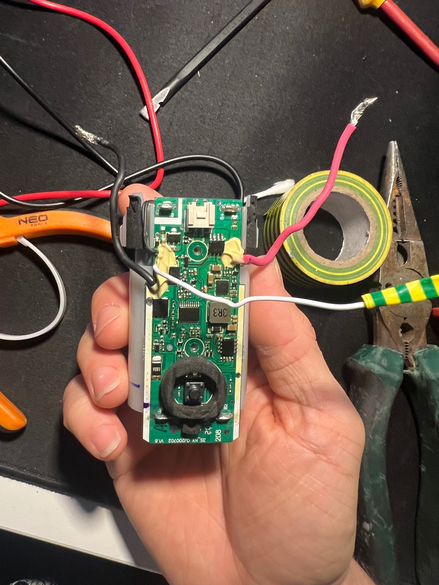 Ręka trzymająca płytkę drukowaną z elementami elektronicznymi, widoczne przewody, taśma izolacyjna i narzędzia, sugerujące naprawę lub modyfikację urządzenia.