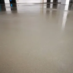 Świeżo wylana, gładka posadzka w jasnym odcieniu szarości z wyraźnym połyskiem, odbijająca fragmenty ścian i filarów w surowym, niedokończonym wnętrzu.