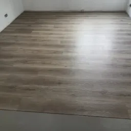 Ułożona podłoga z paneli imitujących drewno w jasnym odcieniu, widoczne gniazdka elektryczne na białej ścianie, fragment surowej podłogi przed panelami.