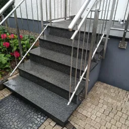 Zewnętrzne schody wejściowe wyłożone ciemnym, nakrapianym granitem, z nowoczesną, metalową balustradą, prowadzące do domu z szarą elewacją i kostką brukową na podłożu.