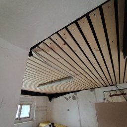 Pogotowie Remontowe Piotr Drozdowski - Montaż drewnianej boazerii na suficie w pomieszczeniu z surowymi ścianami, widoczna lampa sufitowa i okno.