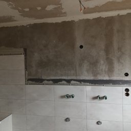 Pogotowie Remontowe Piotr Drozdowski - Ściana w trakcie wykańczania z częściowo położonymi jasnymi płytkami, widoczne otwory na rury i gniazdka, surowy beton na górze ściany i suficie.