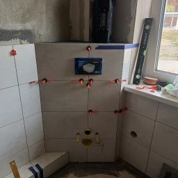 Pogotowie Remontowe Piotr Drozdowski - Ściana łazienki w trakcie układania płytek, widoczne krzyżyki dystansowe, poziomica na parapecie, przygotowane otwory na instalację sanitarną, pudełko po stelażu podtynkowym Geberit.