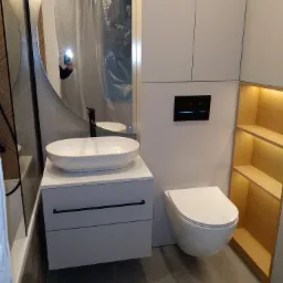 Nowoczesna łazienka z owalną umywalką nablatową, podwieszaną toaletą i drewnianą półką z podświetleniem LED, widoczna osoba robiąca zdjęcie w lustrze.