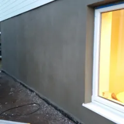 Świeżo otynkowana ściana zewnętrzna budynku z białymi panelami, widoczne okno z zapalonym światłem wewnątrz i fragment drabiny malarskiej.