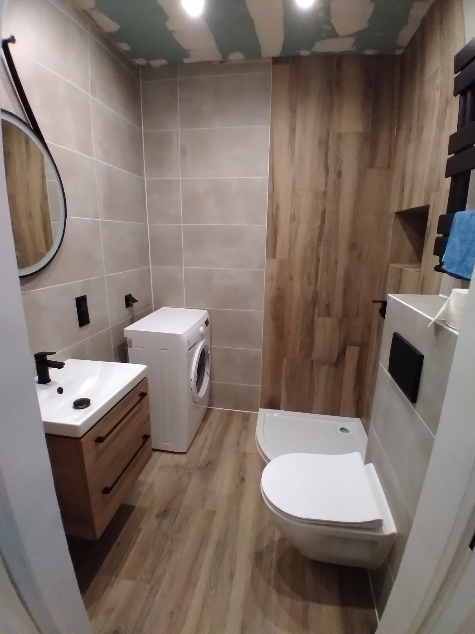 Wykończona łazienka z beżowymi płytkami ściennymi, drewnopodobnymi panelami na podłodze i ścianie, białą umywalką z drewnianą szafką, toaletą podwieszaną i pralką; sufit w trakcie prac...