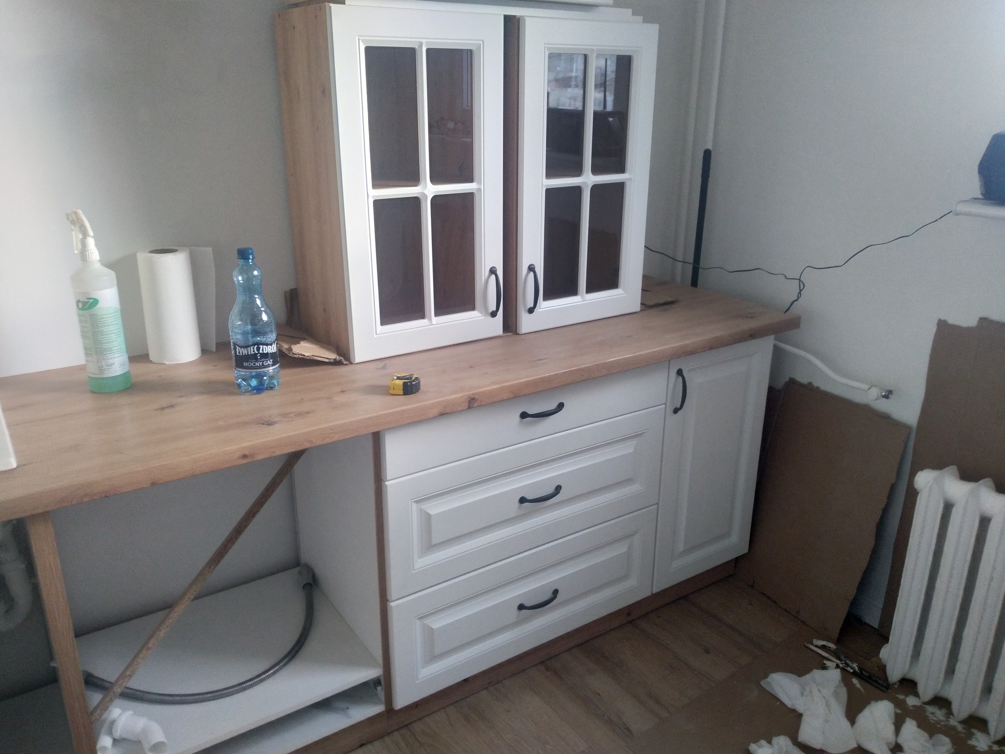Fragment kuchni w trakcie montażu: białe szafki z przeszklonymi frontami, drewniany blat, butelka wody mineralnej i rolka ręcznika papierowego na blacie, kartony i grzejnik obok szafek.