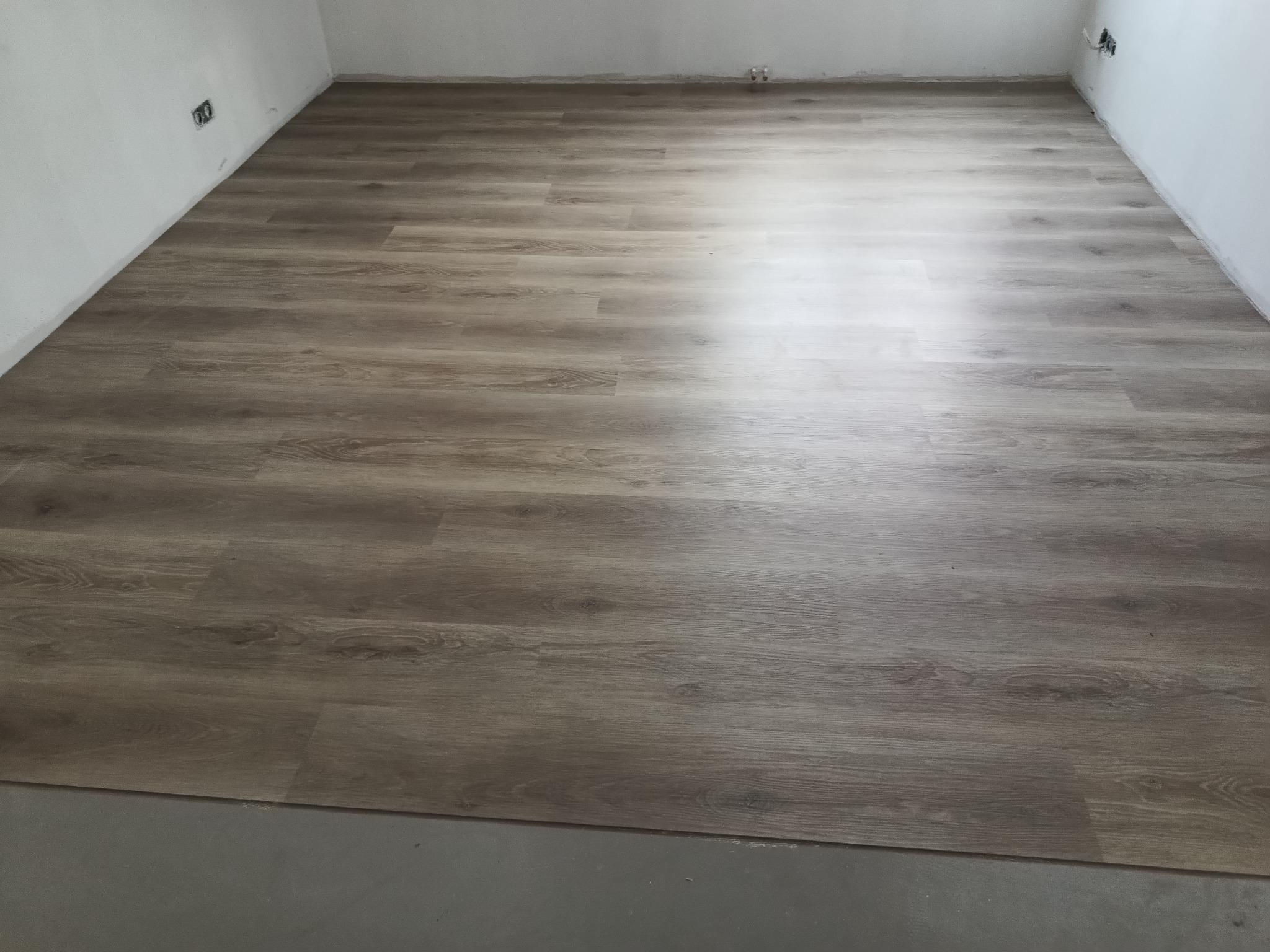 Ułożona podłoga z paneli imitujących drewno w jasnym odcieniu, widoczne gniazdka elektryczne na białej ścianie, fragment surowej podłogi przed panelami.