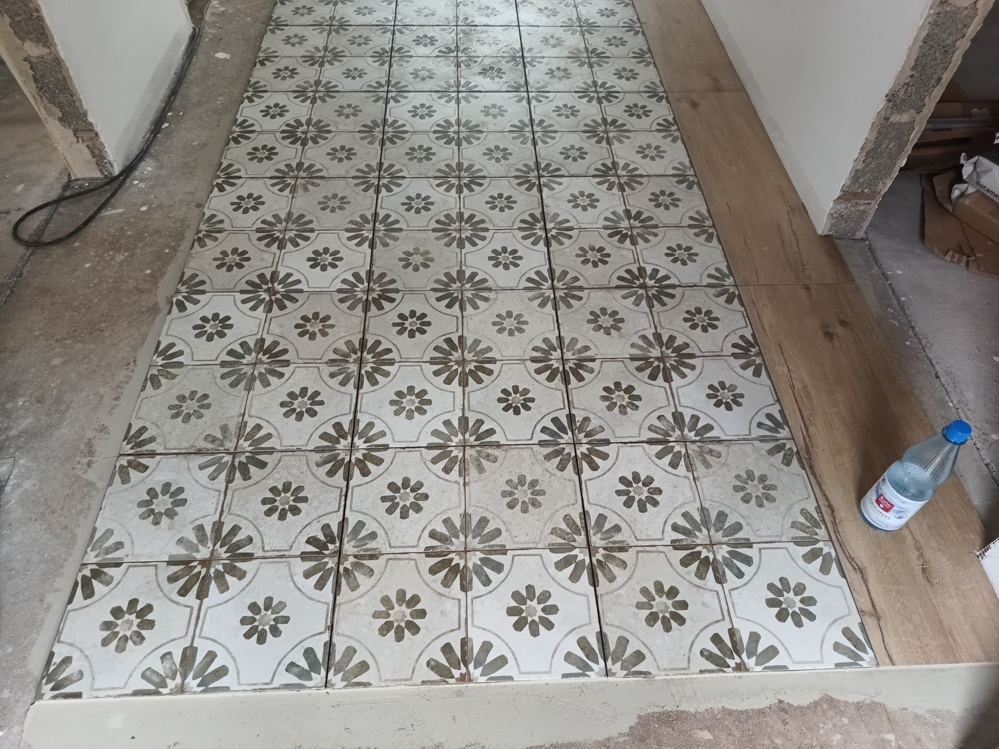 Ułożona podłoga z płytek ceramicznych w kwiatowy wzór, widoczne niedokończone krawędzie i butelka wody mineralnej.