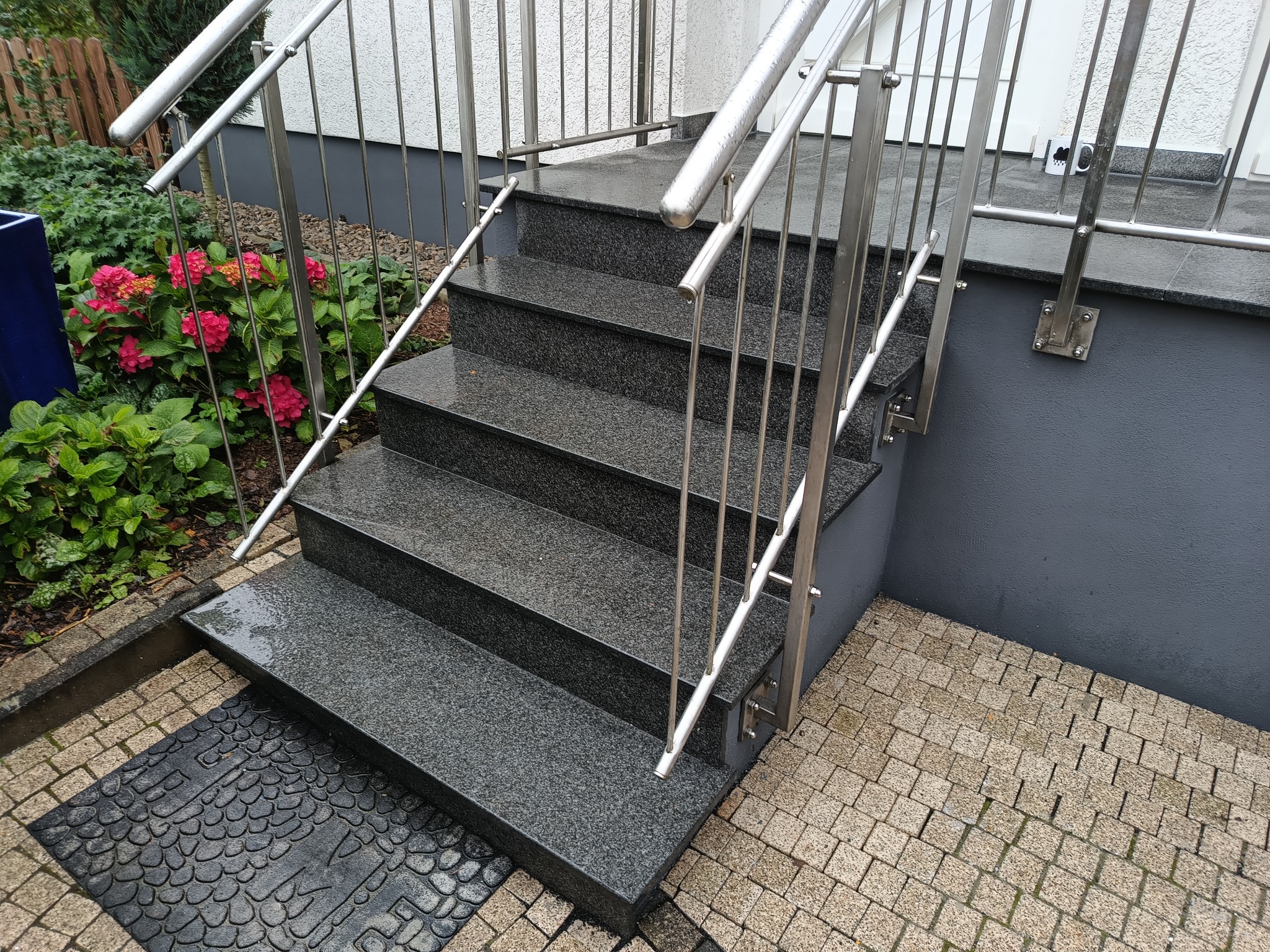 Zewnętrzne schody wejściowe wyłożone ciemnym, nakrapianym granitem, z nowoczesną, metalową balustradą, prowadzące do domu z szarą elewacją i kostką brukową na podłożu.