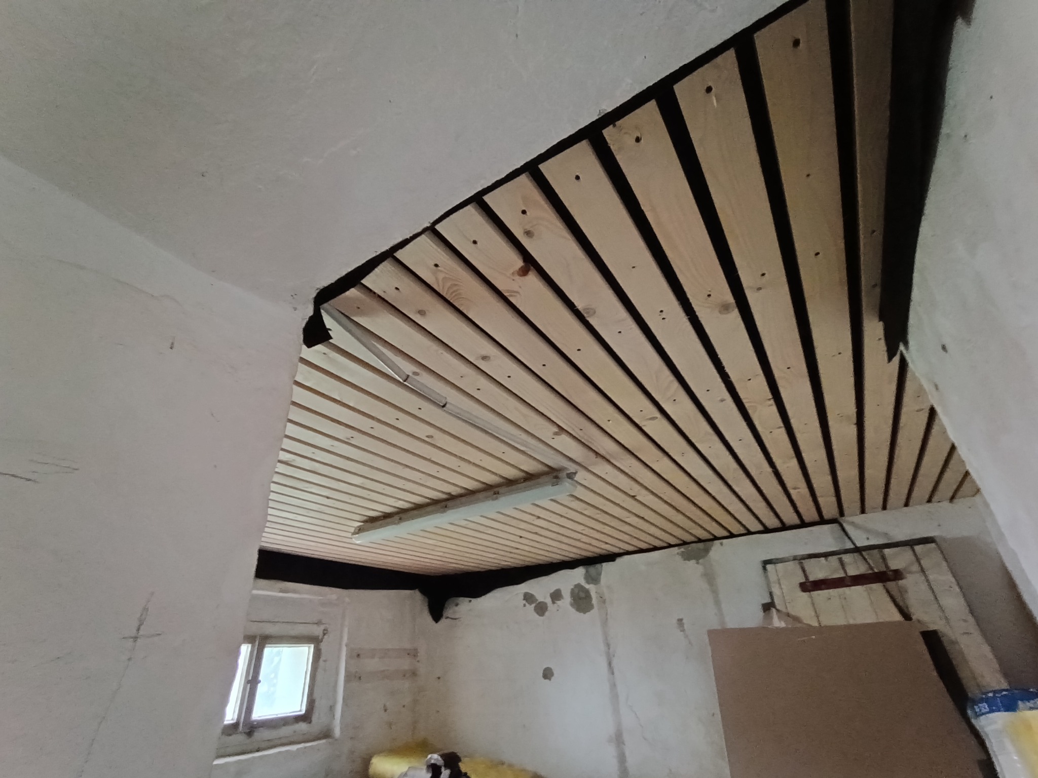 Montaż drewnianej boazerii na suficie w pomieszczeniu z surowymi ścianami, widoczna lampa sufitowa i okno.