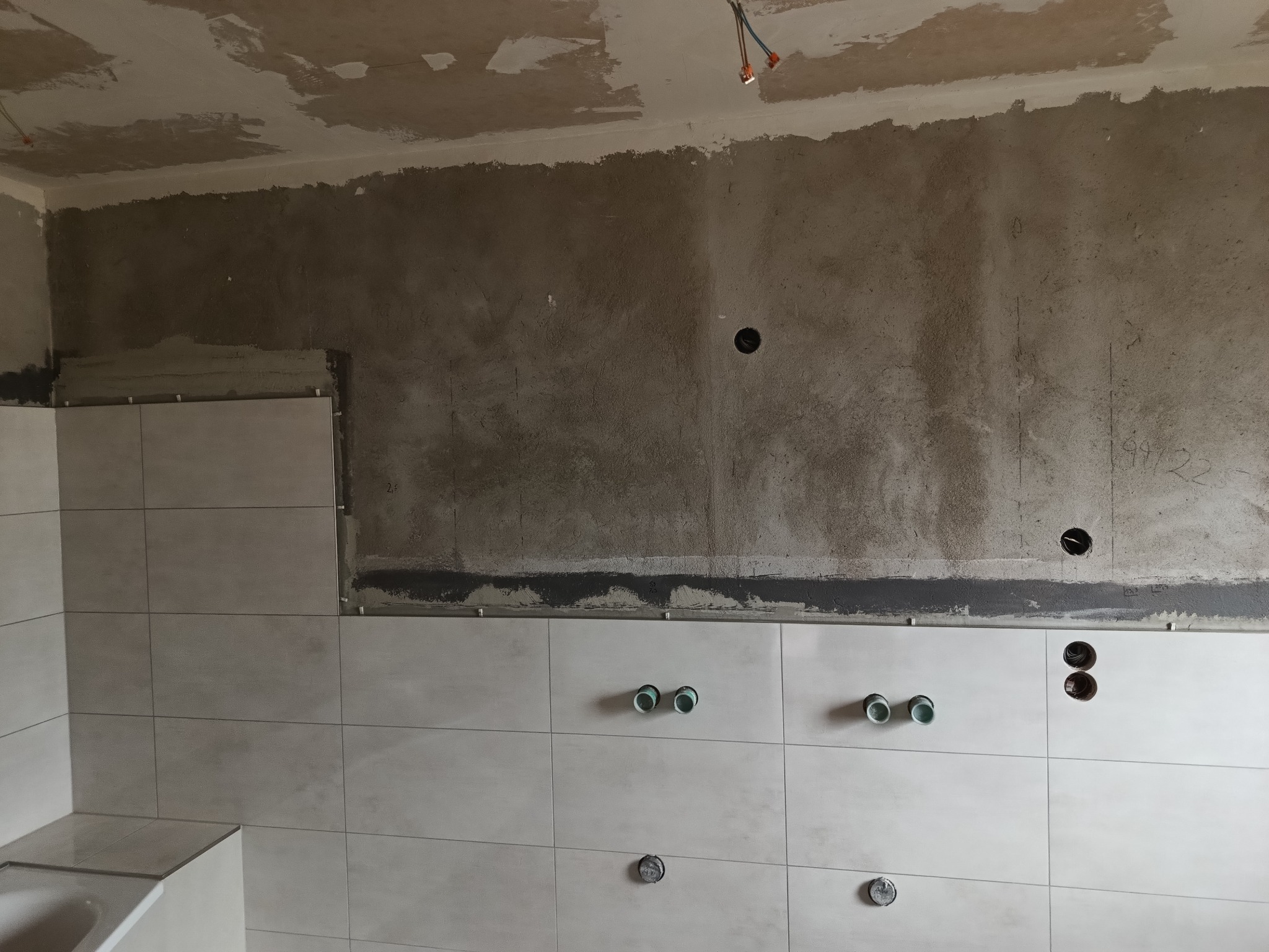 Ściana w trakcie wykańczania z częściowo położonymi jasnymi płytkami, widoczne otwory na rury i gniazdka, surowy beton na górze ściany i suficie.