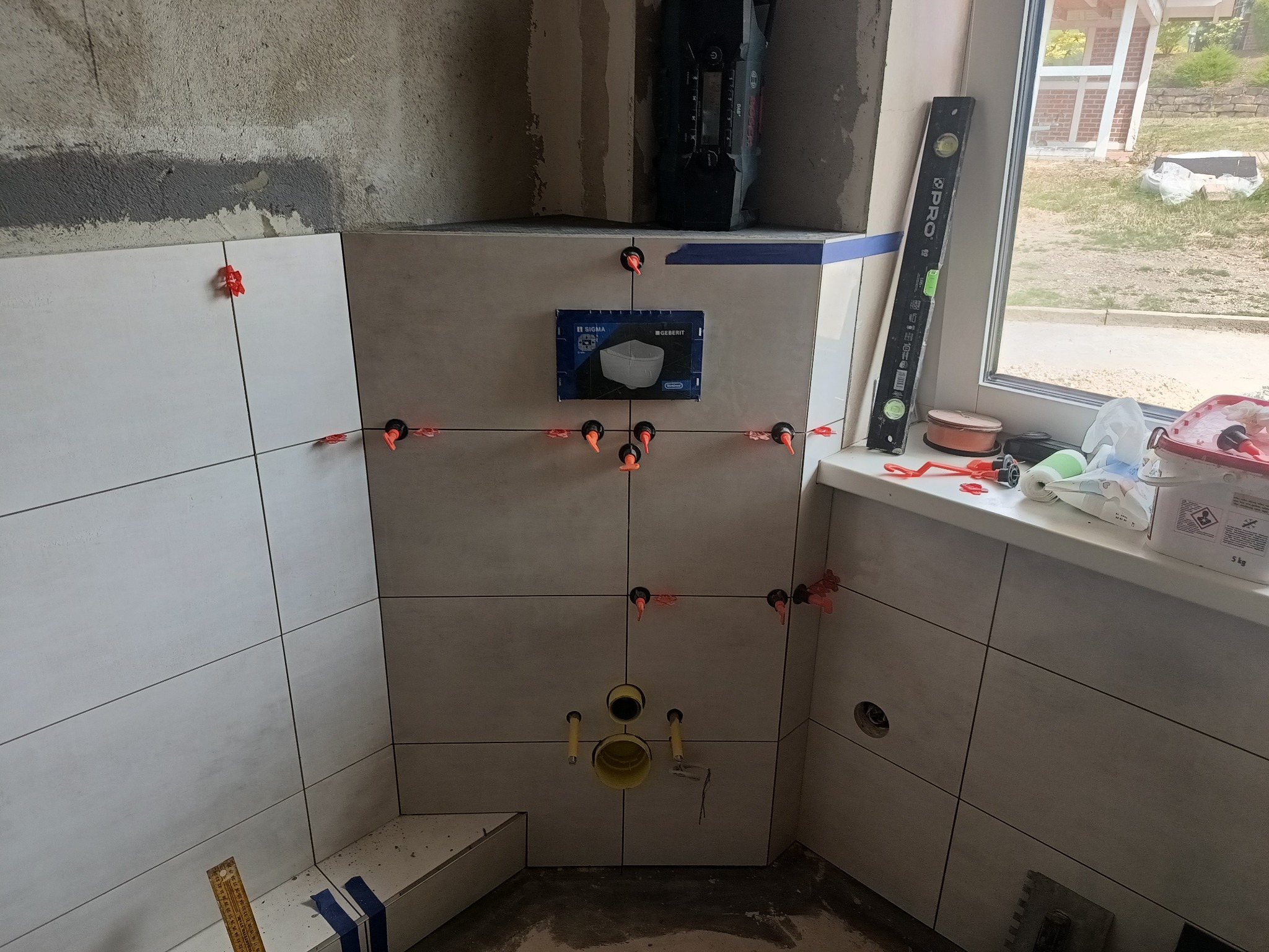 Ściana łazienki w trakcie układania płytek, widoczne krzyżyki dystansowe, poziomica na parapecie, przygotowane otwory na instalację sanitarną, pudełko po stelażu podtynkowym Geberit.