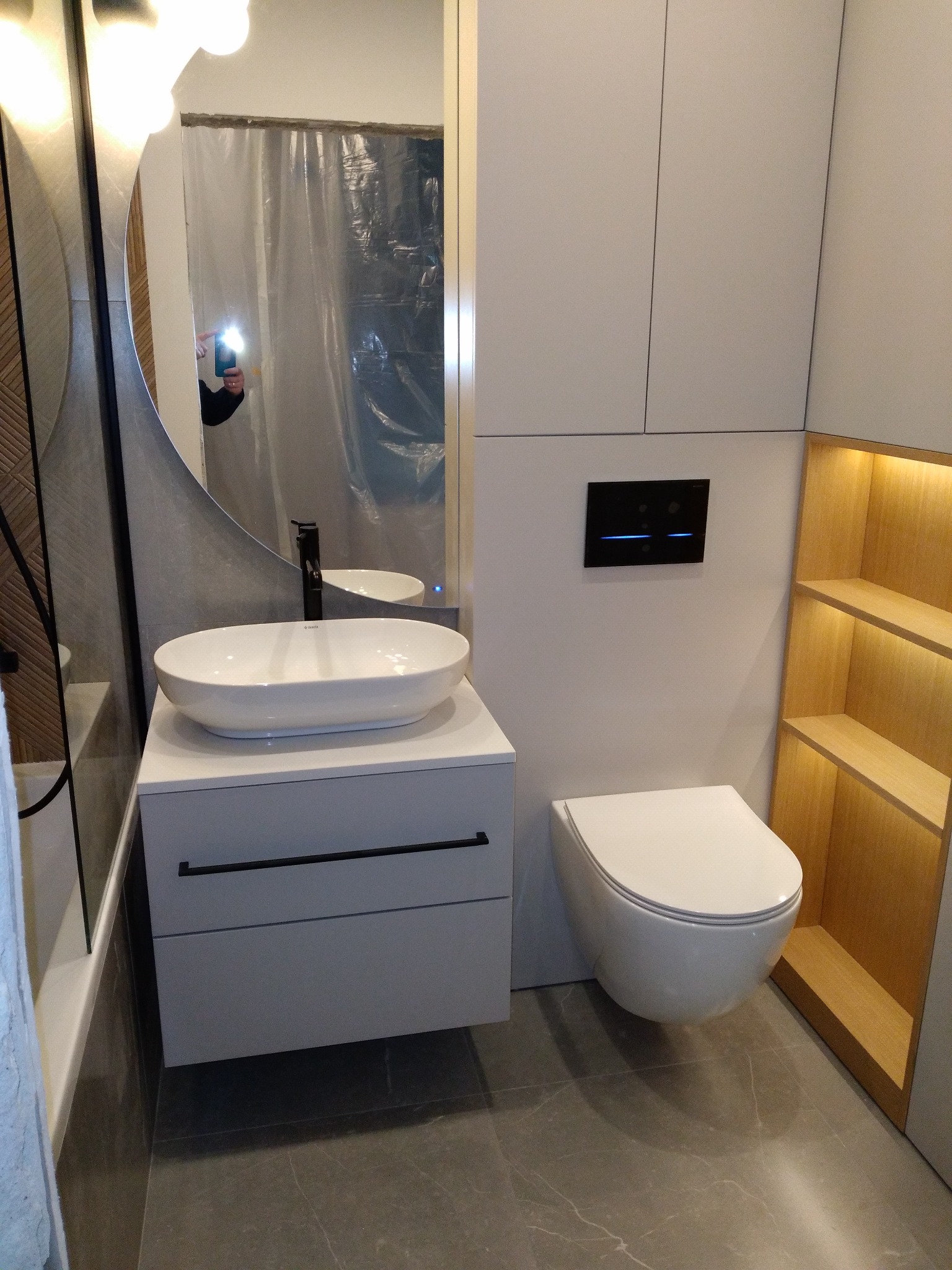 Nowoczesna łazienka z owalną umywalką nablatową, podwieszaną toaletą i drewnianą półką z podświetleniem LED, widoczna osoba robiąca zdjęcie w lustrze.