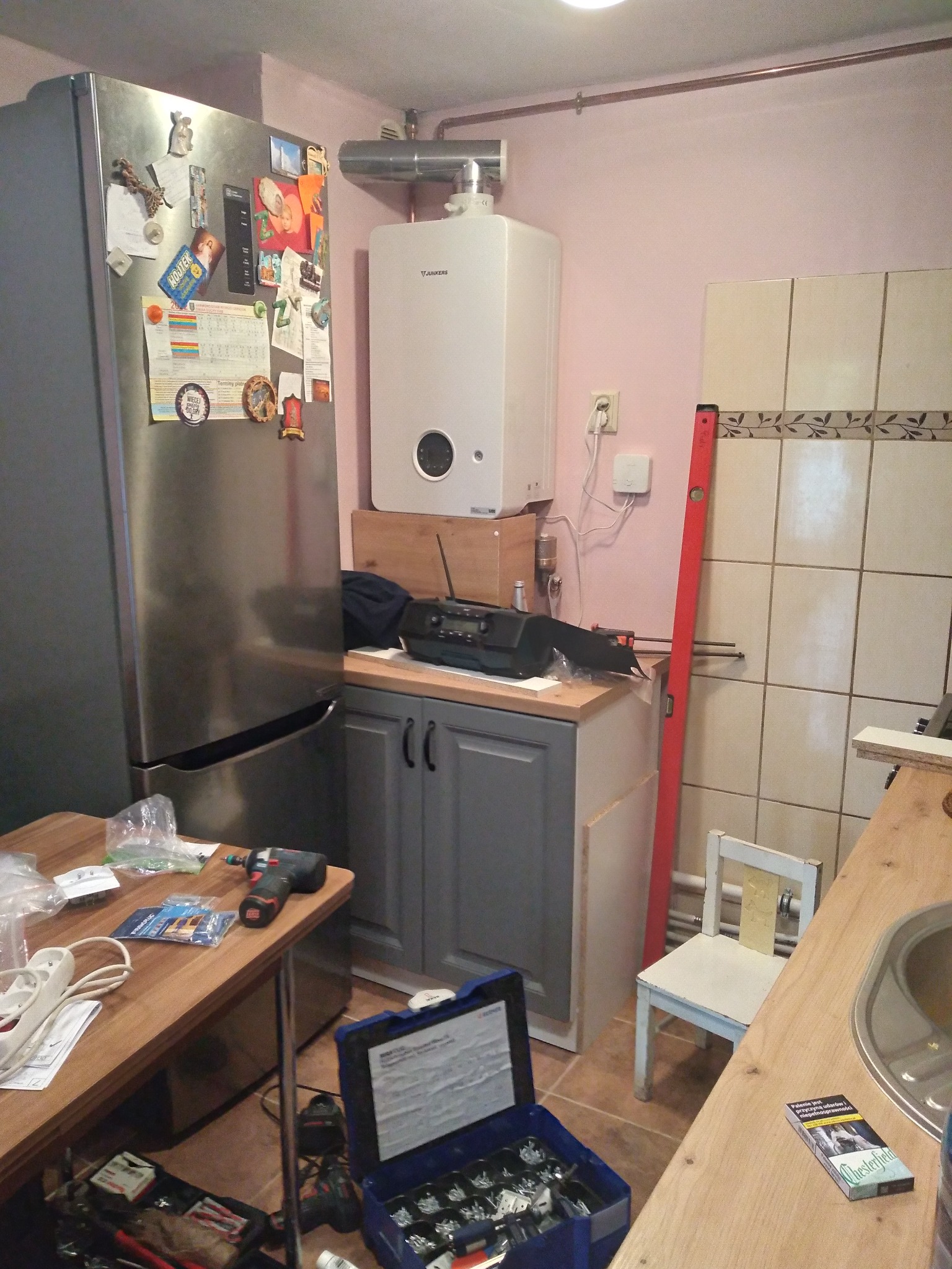 Kocioł gazowy Junkers zamontowany w kuchni, obok lodówki, z widocznymi przewodami, poziomica oparta o ścianę z płytek i narzędzia na stole