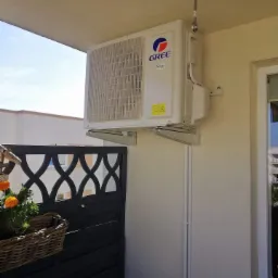 Biała jednostka zewnętrzna klimatyzatora marki Gree zamontowana na balkonie, widoczna instalacja odprowadzająca skropliny, w tle czarna balustrada z dekoracyjnym wzorem i kosz z kwiatami.