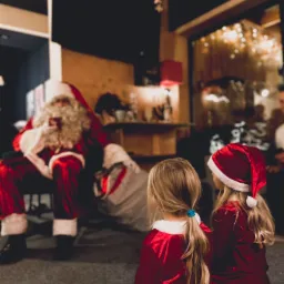 Dwie dziewczynki ubrane w stroje Mikołaja siedzą na podłodze, patrząc na siedzącego Mikołaja w czerwonym stroju z białą brodą w pomieszczeniu z dekoracjami świątecznymi.